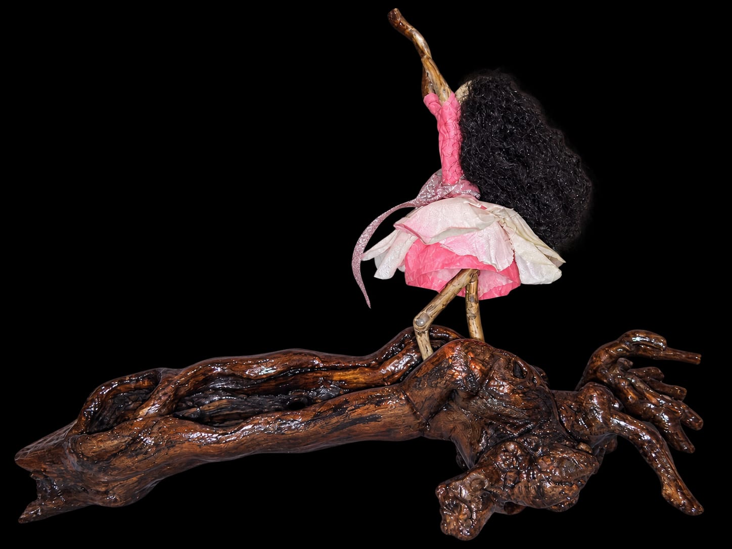 Handmade figure in a pink and white dress with one arm raised on a wooden base